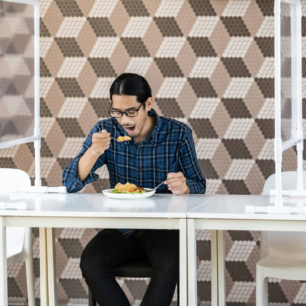 Portrait of asian young adult man eat fried rice food at new normal social distance restaurant with table shiled partition. Restaurant new normal lifestyle and food - Fotó, kép