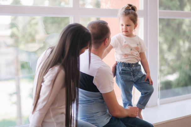Маленькая девочка с мамой и папой сидит у большого окна в студии. Семейная фотосессия - Фото, изображение