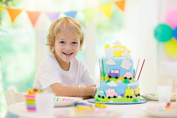 Παιδικό πάρτι γενεθλίων. Παιδική τούρτα με αυτοκίνητο και αεροπλάνο. Παιδί σβήνει κεριά σε πολύχρωμο κέικ. Διακόσμηση πάρτι, σημαίες ουράνιο τόξο, μπαλόνια. Γιορτή θέματος οχήματος. Παιδί γιορτάζει γενέθλια - Φωτογραφία, εικόνα