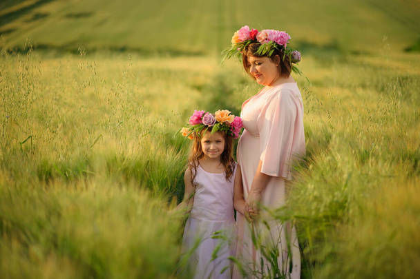 Anya és lánya rózsaszín ruhában egy zöld búzamezőn. A fejükön virágkoszorúk vannak.. - Fotó, kép