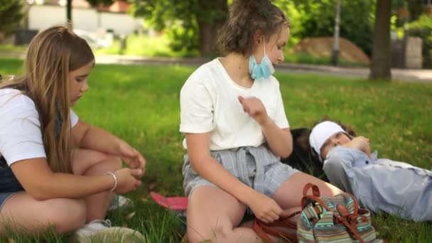 Contactless greeting, new rules. Masked schoolchildren children with backpacks are resting lying on the grass in the park. Post-quarantine life, new normality, back to school - Footage, Video