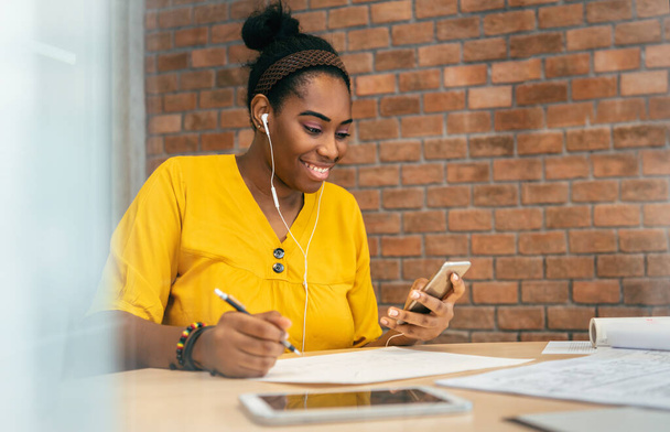 African woman designer using smartphone and earphones for listening to music while working at workplace. African-American Creative Female in Yellow shirt enjoying online video chat with mobile phone - Valokuva, kuva