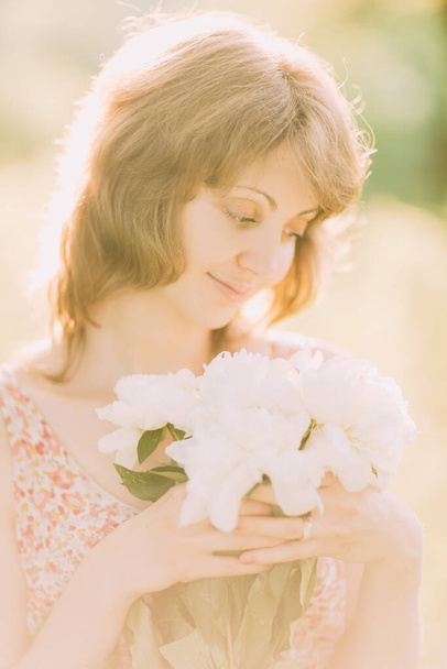 Beautiful young Caucasian blond woman in light dress holding bouquet of white peonies, walking in summer field or garden in sunset. Woman with flowers outdoors - Photo, Image