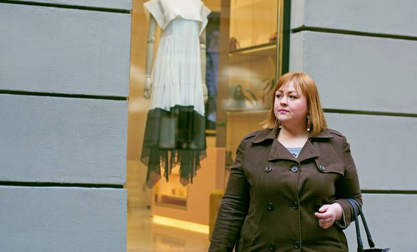 Χαριτωμένο απλή γυναίκα με κόκκινα μαλλιά του μεγέθους συν το μέγεθος βόλτες μέσα από τα ψώνια επιλέγει πράγματα όπως ψώνια και όμορφα κομψά αξεσουάρ αντανακλά στην εμφάνισή της - Φωτογραφία, εικόνα