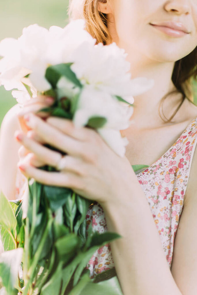Beautiful young Caucasian blond woman in light dress holding bouquet of white peonies, walking in summer field or garden in sunset. Woman with flowers outdoors - Φωτογραφία, εικόνα