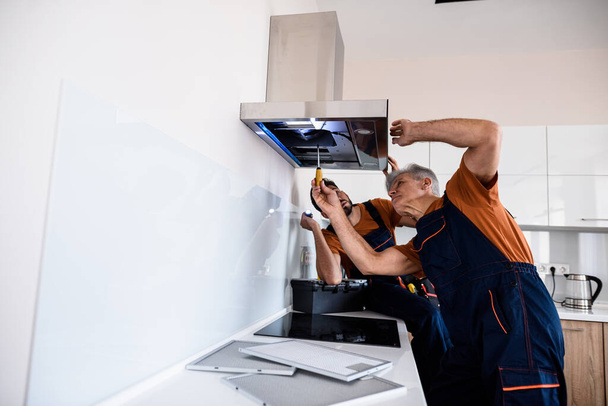 Two workers, handyman in uniform installing or repairing a kitchen extractor, replacing filter in cooker hood. Construction, maintenance and repair concept - 写真・画像