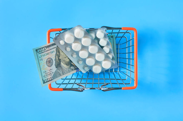 Market basket full of medicaments and banknote of 100 dollars on blue background. Healthcare concept - Фото, зображення