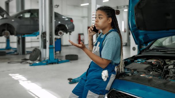 Young african american woman, professional female mechanic talking on phone, leaning on a car with open hood at auto repair shop - Φωτογραφία, εικόνα