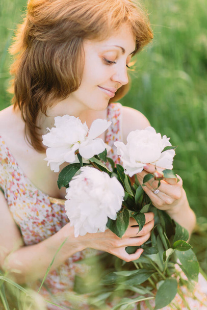 Beautiful young Caucasian blond woman in light dress holding bouquet of white peonies, walking in summer field or garden in sunset. Woman with flowers outdoors - Fotoğraf, Görsel