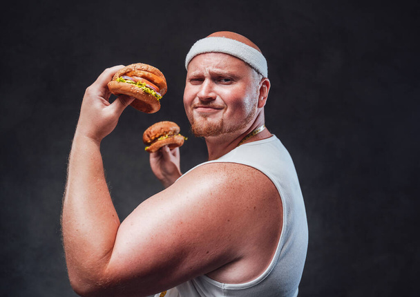 Grappige dikke mannen in het witte verband trekken een gezicht met twee hamburgers in zijn handen - Foto, afbeelding