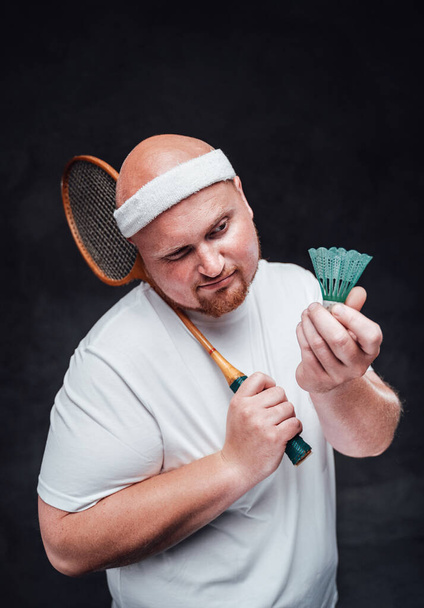 Plešatý tlustý muž s badmintonovou raketou v ruce se na kohouta podívá přimhouřenýma očima. - Fotografie, Obrázek
