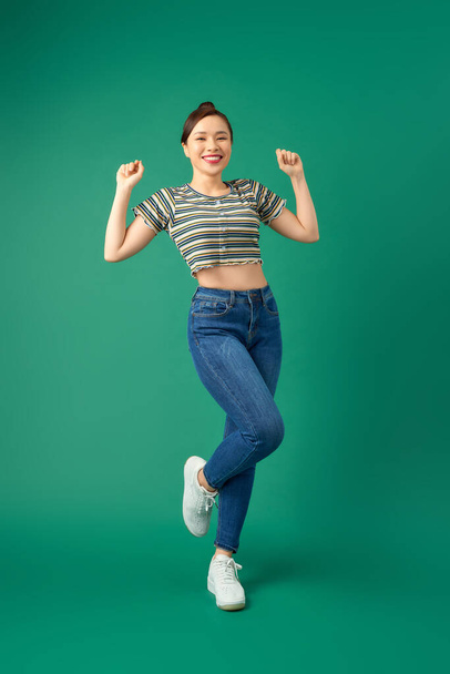 Joyful young Asian woman standing on one leg. Full length view of blissful girl dancing on green background. - Foto, afbeelding