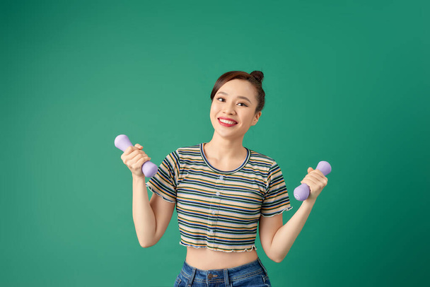 Smiling attractive young Asian woman practicing exercise with dumbell over green background. - Photo, Image