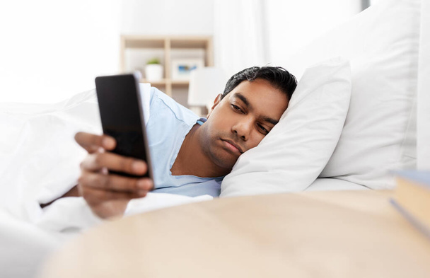 sleepy indian man with smartphone lying in bed - Фото, изображение