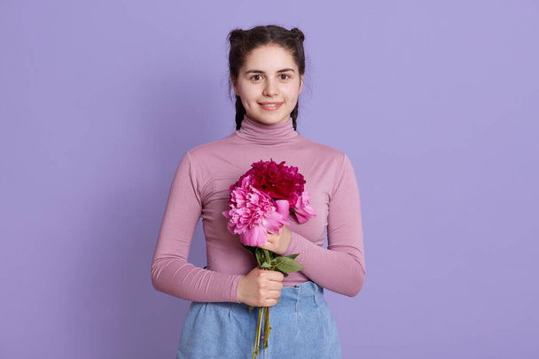 Portrait of happy smiling nice young dark haired girl in casual attire, holding bouquet of peony flowers, standing over lilac background, cute female with pigtails. - Fotó, kép