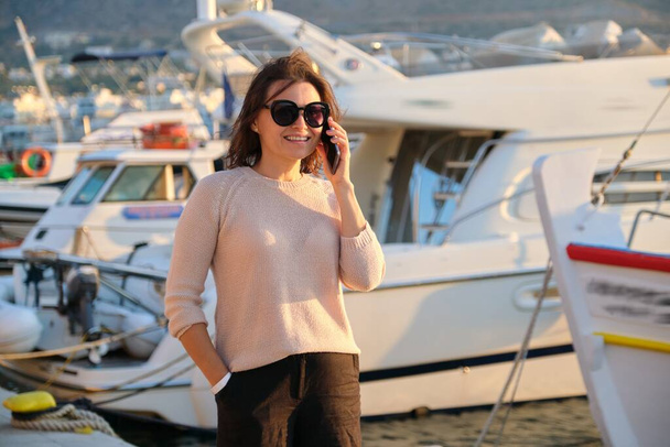Portrait of mature beautiful woman walking in sea pier talking on mobile phone - Photo, image