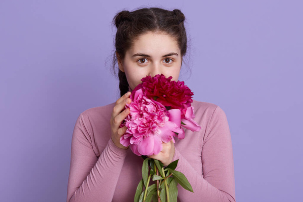 Beauty delicate woman smelling spring flowers and looking directly at camera with charming expression, lady with pigtails holding flowers isolated over lilac wall. - Fotó, kép