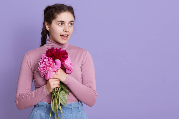 Portrait of brunette young lady wearing casual clothing, embracing bouquet of peonies, female looking aside, standing against lilac background with opened mouth, being astonished. - Foto, afbeelding