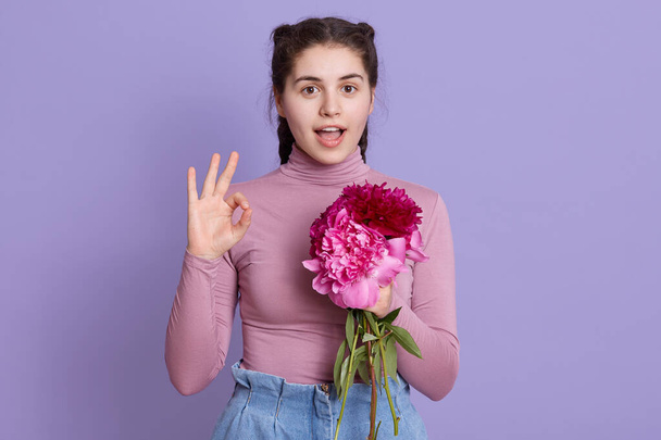 Young beautiful woman wearing casual clothing holding bouquet of roses and burgundy peony flowers, doing ok sign with fingers, excellent symbol, keeping mouth opened. - Foto, immagini