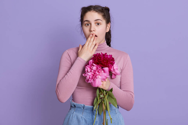 Young woman over lilac wall wearing casual attires folding flowers in hands, being shocked, covering mouth with hand for mistake, posing against lilac wall. - Valokuva, kuva