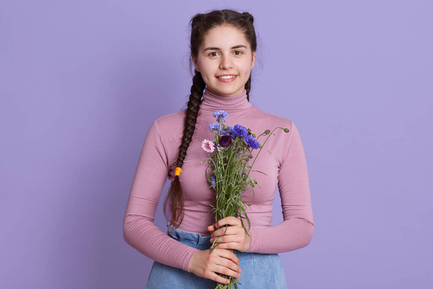 Portrait of gorgeous young brunette woman with long hair and two pigtails, wearing sweater and jeans, holding bouquet of field flowers, standing isolated lilac background. - Fotó, kép