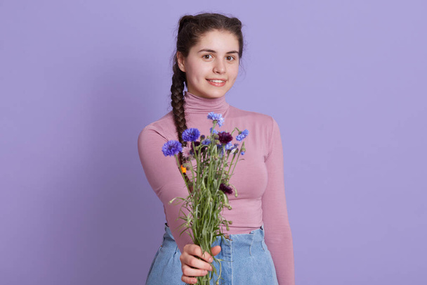 Charming woman holding bouquet of field flowers, female offers flowers to somebody, girl with pigtails posing isolated over lilac background, smiling teenage girl. - Foto, imagen