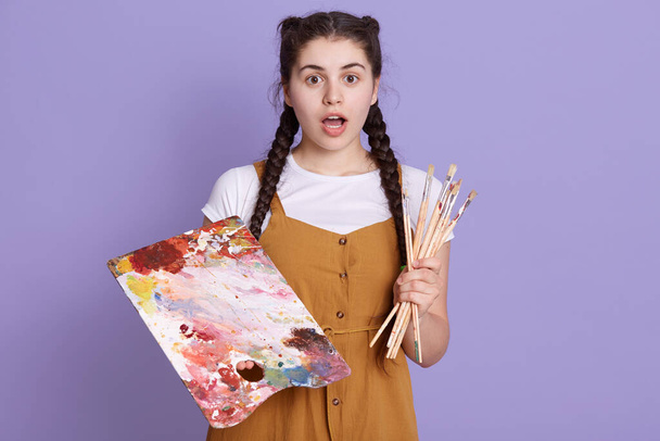 Young talented woman artist with brush and paints posing isolated over lilac background with opened mouth and astonished facial expression, wearing casual attire. - Fotografie, Obrázek