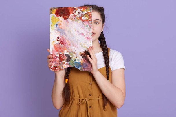 Young woman covering half of her face with easel, looks at camera, wearing casual clothing and standing isolated over lilac background, artist posing with her picture. - Fotografie, Obrázek