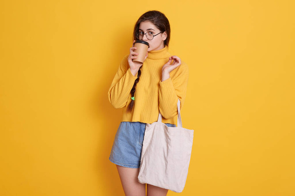 Young woman in stylish clothes with eco bag and drinking hot beverage, enjoying coffee while posing isolated over yellow background, brunette lady with pigtail. - Фото, зображення