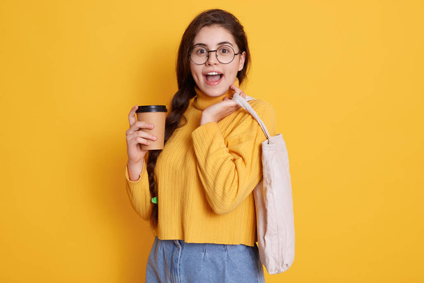 Image of beautiful stylish woman holding eco bag and take away coffee, lady with excited facial expression looking directly at camera, wearing casual clothing and eyewears. - Zdjęcie, obraz