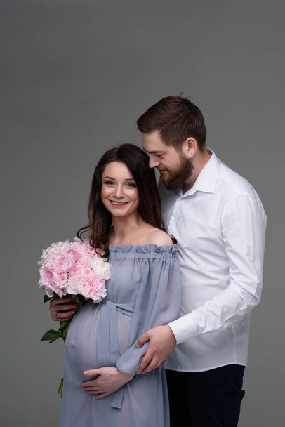 Eine schwangere Frau und ihr Mann umarmen sich vor grauem Hintergrund im Studio. - Foto, Bild