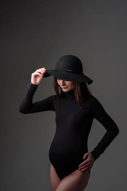 Fiatal gyönyörű terhes nő fekete testöltözékben és kalapban. - Fotó, kép
