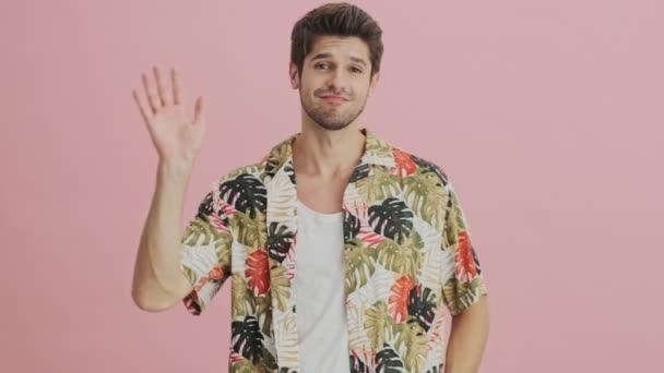 A positive young man is waving his hand to the camera isolated over pink background - Video