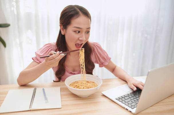 Giovane donna asiatica mangiare tagliatelle e lavorare con il computer portatile a casa. - Foto, immagini