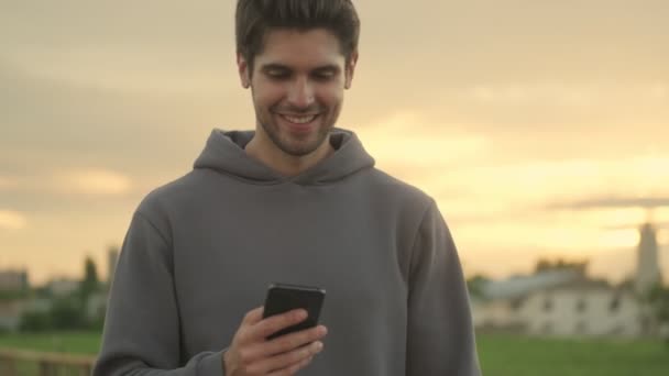 Un jeune homme souriant utilise son smartphone en marchant dehors le matin - Séquence, vidéo