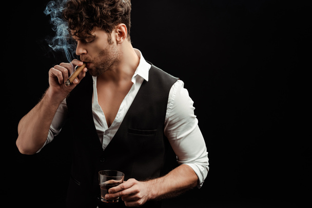 Handsome man smoking cigar and holding glass of whiskey isolated on black  - Foto, imagen