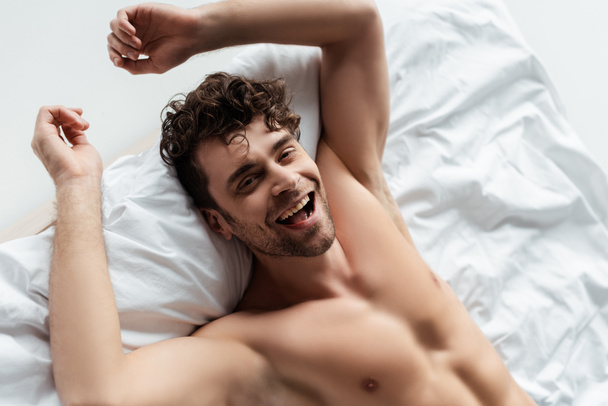 High angle view of cheerful shirtless man smiling at camera on bed isolated on white - Photo, Image
