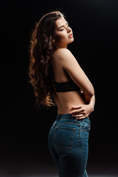 Beautiful brunette woman in bra and jeans with closed eyes isolated on black  - Photo, Image