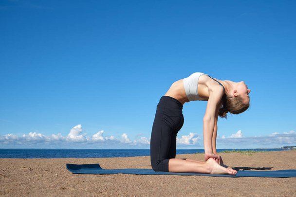 Güneşli bir sabahta kumsalda yoga yapan sıska bir kız spor yapmadan önce esneme hareketleri yapıyor. - Fotoğraf, Görsel