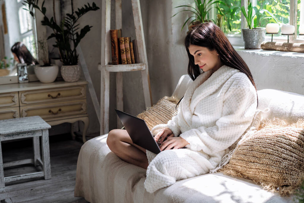 Side view of young adult woman sitting on sofa in bathrobe, using modern laptop computer. girl making smiling face, resting on weekend at home - Foto, Bild