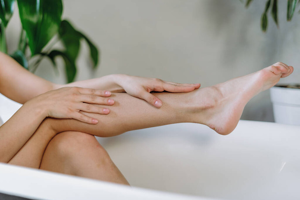 Huidverzorgingsconcept. Gewassen uitzicht op jonge volwassen vrouw liggend in bad, aanraken van haar zachte huid op perfecte benen, brengen de ochtend en rusten in de badkamer - Foto, afbeelding