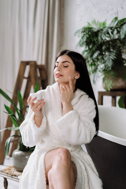 Vertical photo of young adult woman in bathrobe, apply face cream on skin, using skincare and bodycare cosmetic product with natural ingredients. Girl spending morning at bathroom, sitting on chair - Фото, зображення