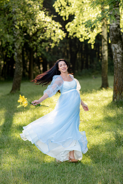 Pregnant woman posing in a blue dress on a background of green trees. - Фото, изображение