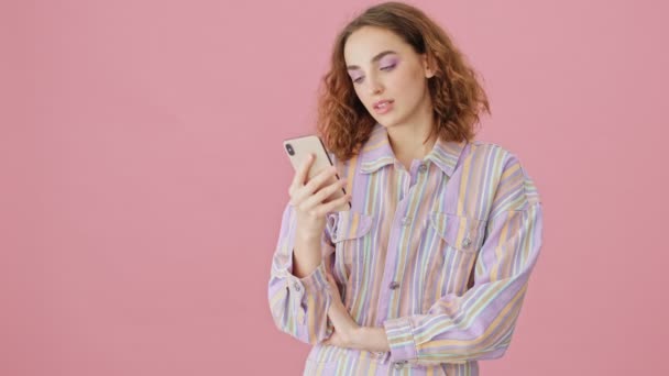 A beautiful young woman is using a smartphone standing isolated over pink background in studio - Imágenes, Vídeo