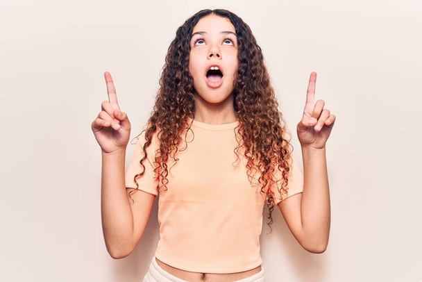 Beautiful kid girl with curly hair wearing casual clothes amazed and surprised looking up and pointing with fingers and raised arms.  - Photo, Image
