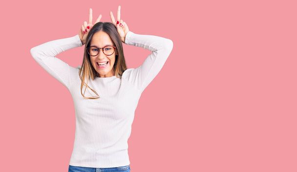 Beautiful brunette young woman wearing casual white sweater and glasses posing funny and crazy with fingers on head as bunny ears, smiling cheerful  - Photo, Image