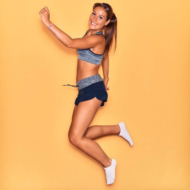 Young beautiful hispanic sportswoman wearing sporty clothes smiling happy. Jumping with smile on face over isolated yellow background. - Foto, afbeelding