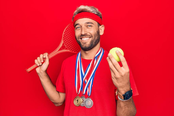 Guapo deportista rubio con barba ganando medallas jugando al tenis usando raqueta y pelota con aspecto positivo y feliz de pie y sonriendo con una sonrisa confiada mostrando los dientes
 - Foto, Imagen