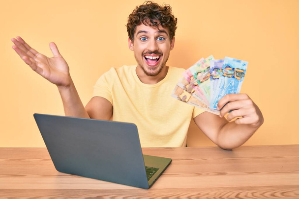 Młody biały mężczyzna z kręconymi włosami siedzi na stole pracując z laptopem i trzymając kanadyjskie dolary banknoty świętując zwycięstwo szczęśliwym uśmiechem i zwycięską ekspresją z podniesionymi rękami  - Zdjęcie, obraz