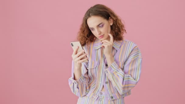A serious young woman is using her smartphone standing isolated over pink background in studio - Imágenes, Vídeo